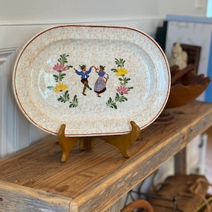 May include: An oval ceramic platter with a speckled beige and white background. The center features a colorful illustration of two figures dancing, surrounded by a decorative wreath of green leaves, pink flowers, and yellow flowers.