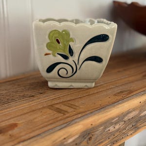 May include: A square ceramic planter with a scalloped top edge and a crackled off-white glaze. The front features a painted floral design in shades of light green, dark blue, and a small touch of red, with a base that has a notched edge detail.