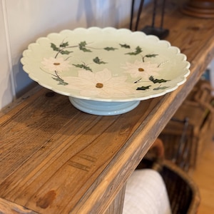 May include: A pale green pedestal cake stand with a scalloped edge, featuring a hand-painted design of white poinsettia flowers and dark green holly leaves arranged in a circular pattern around the center of the plate.