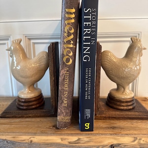 May include: A decorative bookshelf arrangement featuring two ceramic rooster figurines flanking a pair of books. The books are titled "Mexico: A History in Art" and "Stories in Sterling". The rooster figurines are beige and sit on wooden bases.
