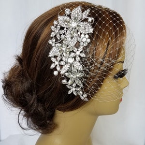 Puede incluir: Un peine de pelo de novia plateado con un dise&ntilde;o floral con brillantes transparentes. El peine se lleva en un maniqu&iacute; con un mo&ntilde;o marr&oacute;n y un velo de jaula de p&aacute;jaros blanco.