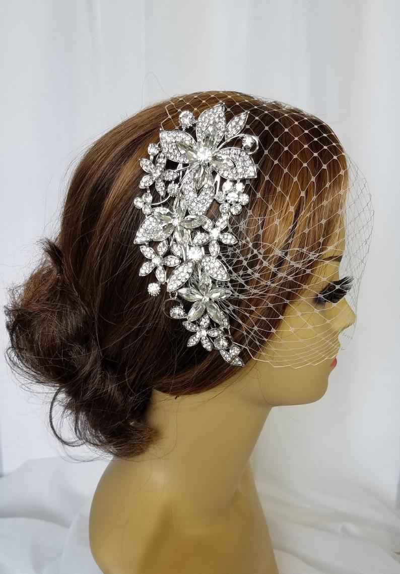 Puede incluir: Un peine de pelo de novia plateado con un dise&ntilde;o floral con brillantes transparentes. El peine se lleva en un maniqu&iacute; con un mo&ntilde;o marr&oacute;n y un velo de jaula de p&aacute;jaros blanco.