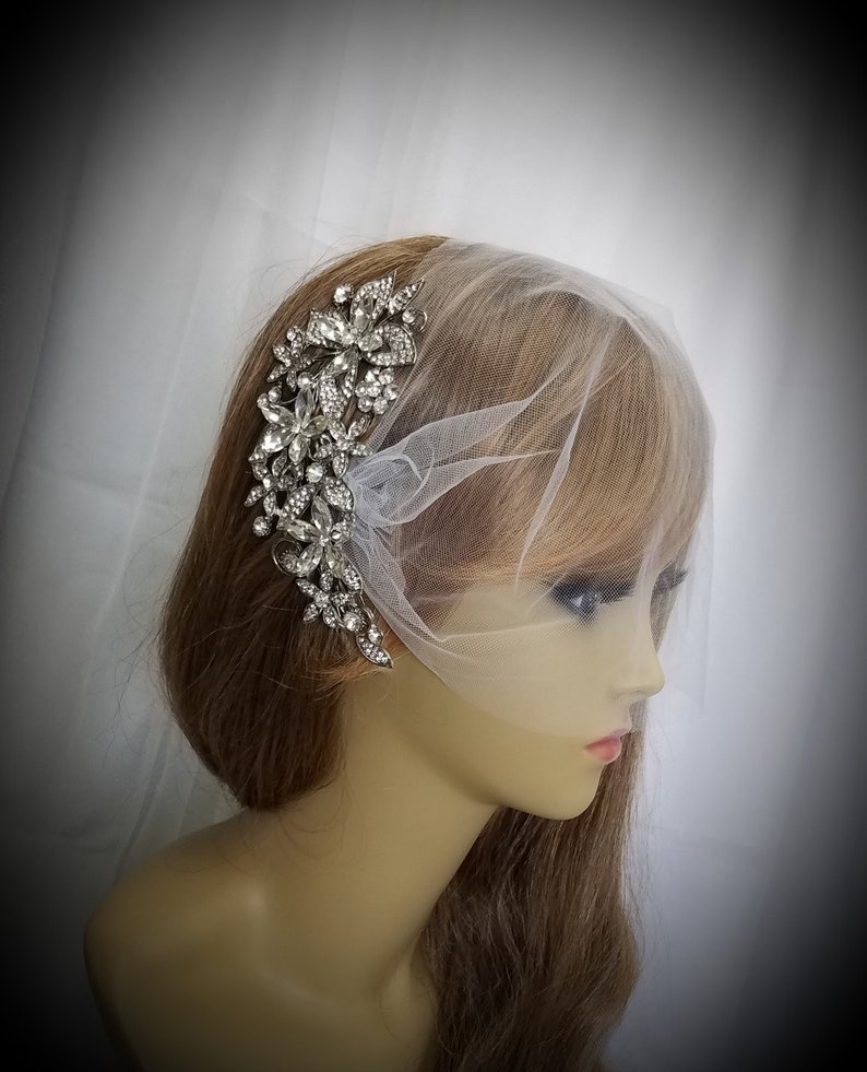 Puede incluir: Un peine de pelo de novia plateado con un dise&ntilde;o floral con brillantes de cristal transparentes. El peine se lleva en el pelo de un maniqu&iacute; con un velo de tul blanco.