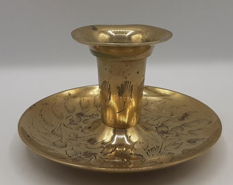 May include: A gold metal candlestick with an ornate design. The candlestick has a round base and a tall, cylindrical holder.