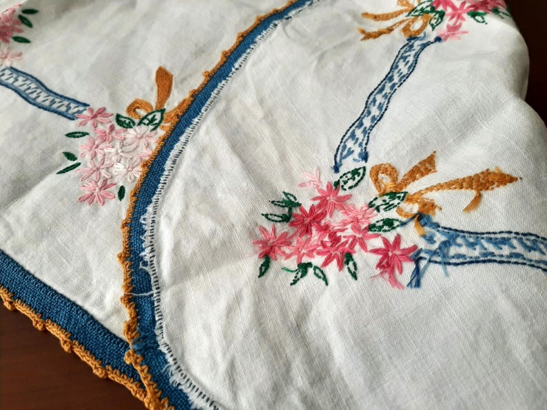 Puede incluir: Tela de lino blanco con dise&ntilde;os florales y de cintas bordadas. El bordado presenta flores rosas, hojas verdes y detalles de cintas azules y doradas. Los bordes est&aacute;n ribeteados con un borde de ganchillo azul y dorado.