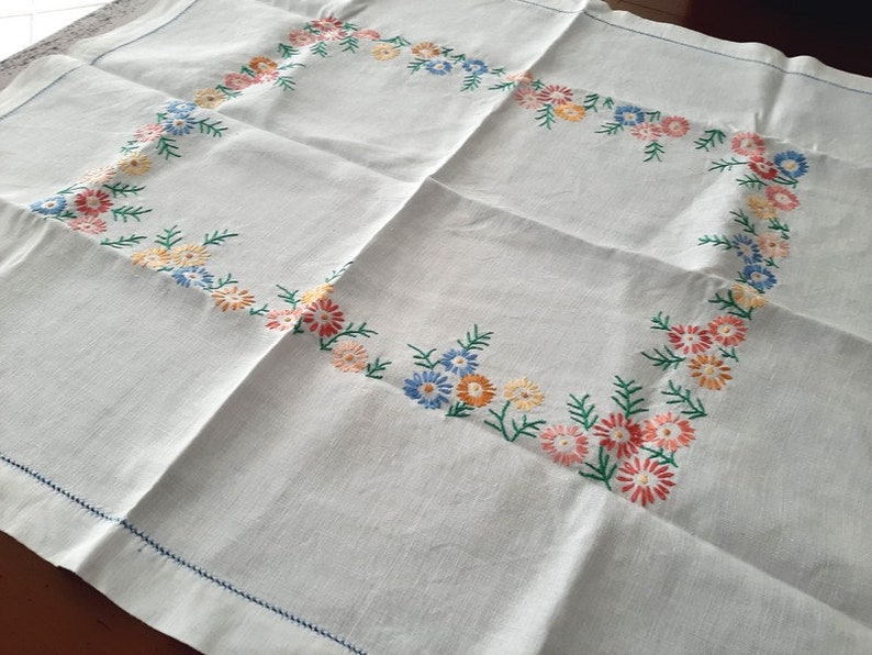 May include: White linen tablecloth with a delicate embroidered border of colorful flowers. The flowers are in shades of blue, pink, yellow, and green.