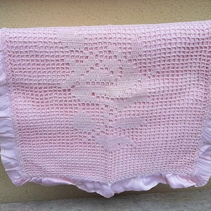 Peut inclure: Une couverture de bébé en crochet rose avec un motif en crochet blanc au centre. La couverture a un bord festonné.