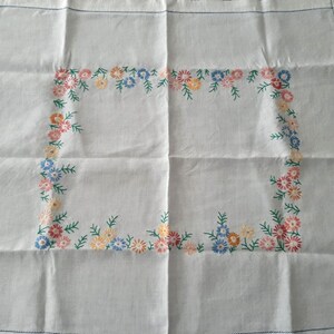 May include: White linen tablecloth with a floral embroidered border. The border features small pink, yellow, and blue flowers with green leaves.