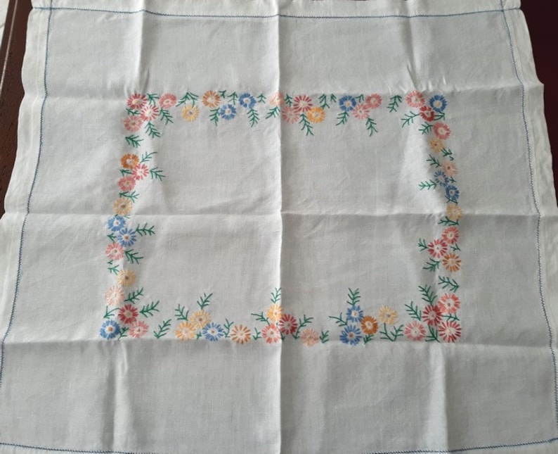 May include: White linen tablecloth with a floral embroidered border. The border features small pink, yellow, and blue flowers with green leaves.