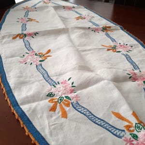 Puede incluir: Camino de mesa ovalado blanco con bordados de flores rosas, lazos naranjas y detalles de cinta azul. Los bordes est&aacute;n adornados con ganchillo azul y naranja. Un textil decorativo para el comedor.