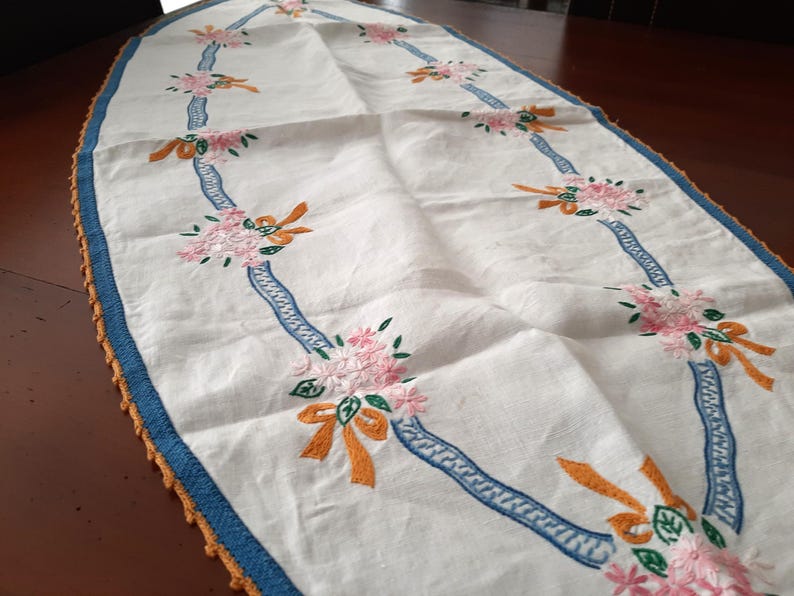 Puede incluir: Camino de mesa ovalado blanco con bordados de flores rosas, lazos naranjas y detalles de cinta azul. Los bordes est&aacute;n adornados con ganchillo azul y naranja. Un textil decorativo para el comedor.