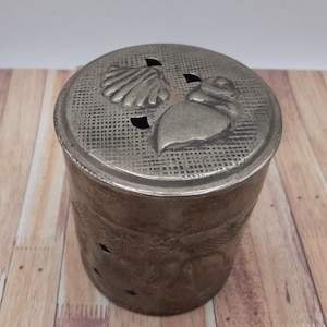 May include: A silver metal container with a lid featuring a seashell design. The container has a textured surface and several small holes.