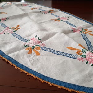 Puede incluir: Camino de mesa ovalado blanco con bordados de flores rosas, lazos naranjas y detalles de cinta azul. Los bordes est&aacute;n ribeteados con un borde de ganchillo azul y naranja. Una pieza decorativa para una mesa.