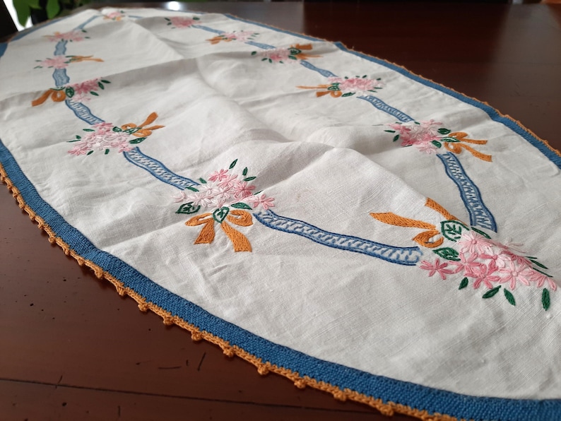 Puede incluir: Camino de mesa ovalado blanco con bordados de flores rosas, lazos naranjas y detalles de cinta azul. Los bordes est&aacute;n ribeteados con un borde de ganchillo azul y naranja. Una pieza decorativa para una mesa.