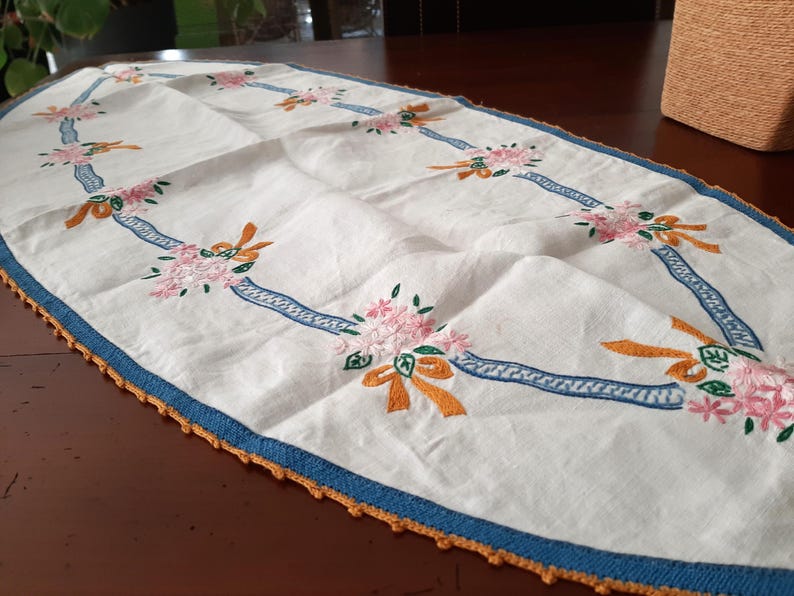Puede incluir: Camino de mesa ovalado blanco con dise&ntilde;o floral y de cintas bordadas. El camino de mesa presenta flores rosas, lazos naranjas y detalles de cinta azul. Los bordes est&aacute;n ribeteados con un borde azul y ganchillo naranja.