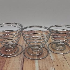 May include: Three silver metal egg holders in a spiral design. The holders are on a wooden surface.