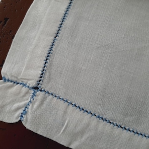 May include: A white linen napkin with a blue embroidered border. The napkin has a scalloped edge.