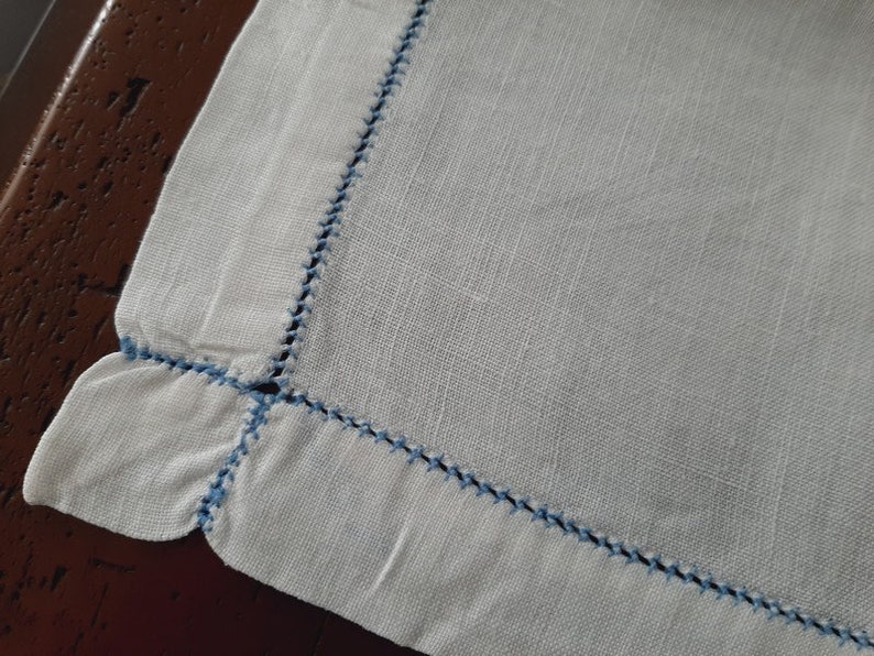 May include: A white linen napkin with a blue embroidered border. The napkin has a scalloped edge.