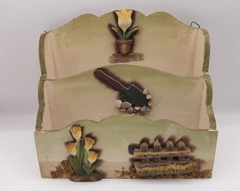May include: A wooden wall-mounted mail holder with a green and cream paint finish. It features three compartments and decorative details of flowers, a shovel, and a fence.