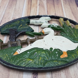 Peut inclure: Un dessous de plat en fonte avec une oie blanche et un motif d'arbre vert, de clôture et d'herbe.