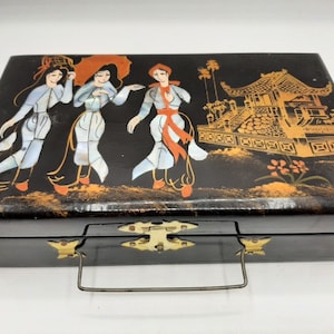 Può includere: Una scatola di legno nero con una fibbia e una maniglia dorate. Il coperchio è decorato con una scena dipinta di tre donne in abiti tradizionali, un pagoda e fiori. Le donne sono dipinte in tonalità di bianco, rosso e blu.