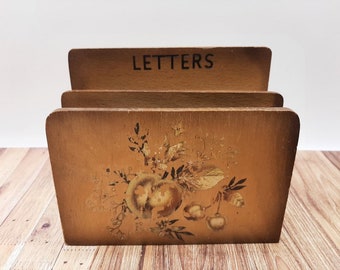 May include: A wooden letter holder with the word "LETTERS" printed on the top. The holder has a floral design with gold accents and is painted in shades of brown.