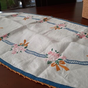 Puede incluir: Camino de mesa blanco bordado con ramos de flores rosas, lazos naranjas y detalles de cinta azul. El camino de mesa tiene un borde azul con un borde de ganchillo naranja. Una pieza decorativa para una mesa.