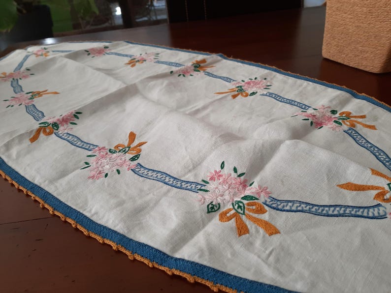 Puede incluir: Camino de mesa blanco bordado con ramos de flores rosas, lazos naranjas y detalles de cinta azul. El camino de mesa tiene un borde azul con un borde de ganchillo naranja. Una pieza decorativa para una mesa.