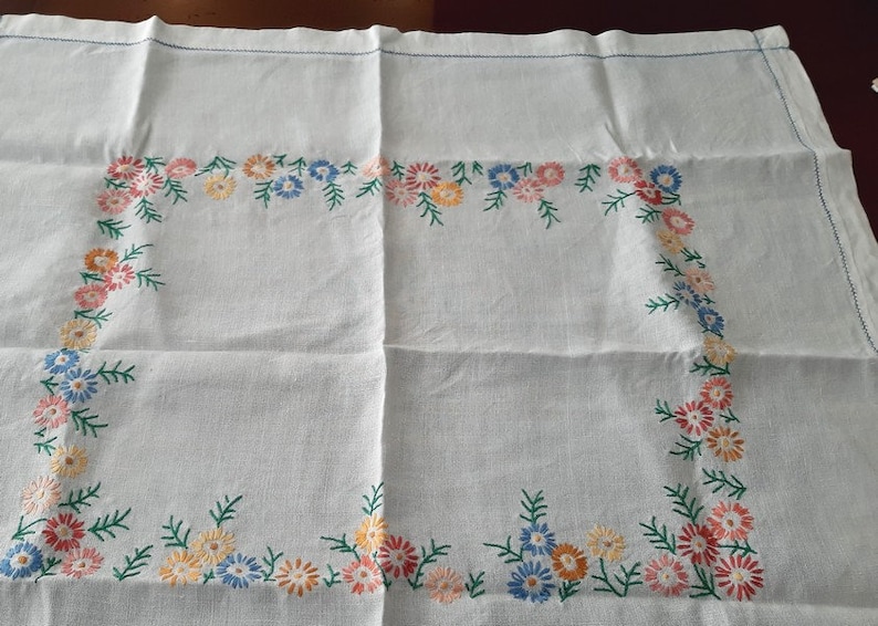 May include: White linen tablecloth with a delicate floral embroidery design in shades of pink, blue, and yellow. The embroidery features a repeating pattern of small flowers and leaves around the border.