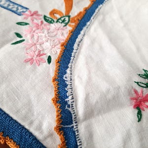 Puede incluir: Primer plano de un textil de lino blanco con detalles florales bordados. Se presentan flores rosas con hojas verdes, junto con adornos azules y naranjas. Los bordes est&aacute;n acabados con una puntada decorativa.