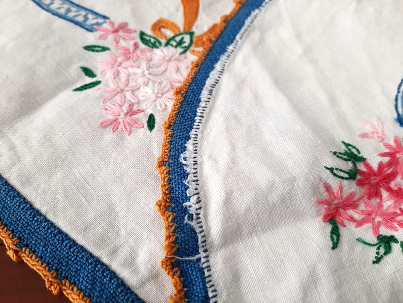 Puede incluir: Primer plano de un textil de lino blanco con detalles florales bordados. Se presentan flores rosas con hojas verdes, junto con adornos azules y naranjas. Los bordes est&aacute;n acabados con una puntada decorativa.
