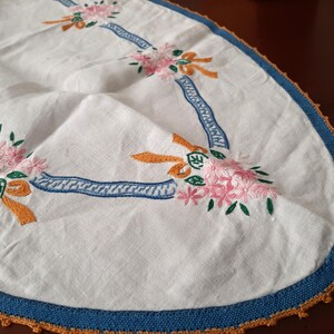 Puede incluir: Mantel ovalado blanco con flores rosas bordadas, lazos naranjas y detalles de cinta azul. El borde est&aacute; adornado con un borde azul y ganchillo naranja. Un textil decorativo vintage.