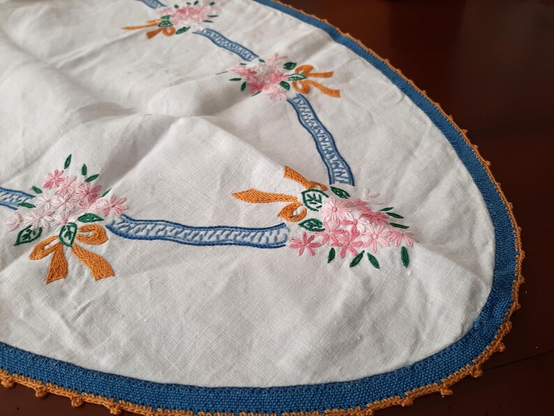 Puede incluir: Mantel ovalado blanco con flores rosas bordadas, lazos naranjas y detalles de cinta azul. El borde est&aacute; adornado con un borde azul y ganchillo naranja. Un textil decorativo vintage.