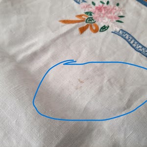 Puede incluir: Pa&ntilde;o de lino blanco con detalles bordados de flores y cintas en rosa, naranja y verde. Se ve un borde decorativo azul a lo largo del borde. El pa&ntilde;o tiene algunas manchas marrones.