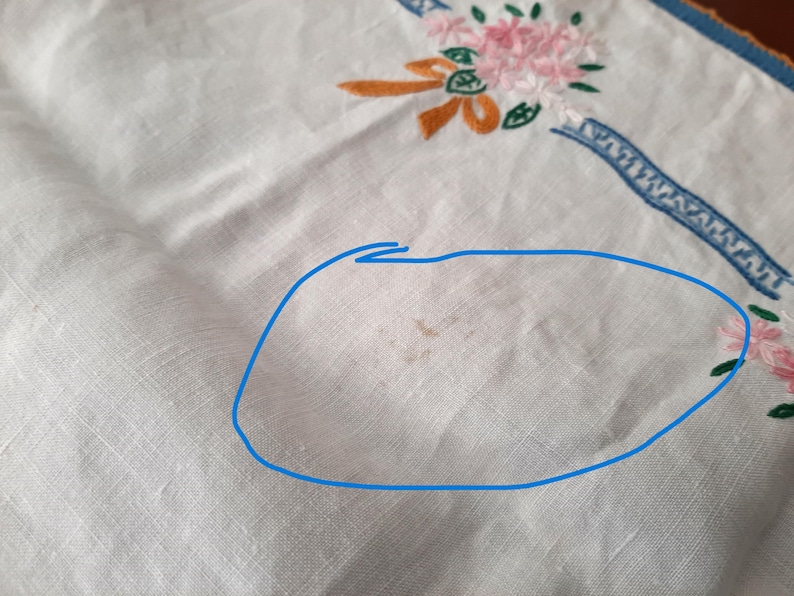 Puede incluir: Pa&ntilde;o de lino blanco con detalles bordados de flores y cintas en rosa, naranja y verde. Se ve un borde decorativo azul a lo largo del borde. El pa&ntilde;o tiene algunas manchas marrones.