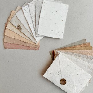 Könnte beinhalten: Eine Sammlung handgefertigter Papiere und Umschläge in verschiedenen neutralen Farbtönen. Das Papier hat eine strukturierte, organische Optik mit sichtbaren Fasern und Einschlüssen. Einige Blätter weisen eingebettete Blätter auf. Die Umschläge sind mit einem dekorativen Wachssiegel verschlossen.