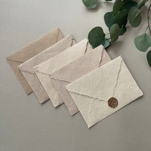 Puede incluir: Cinco sobres en varios tonos de beige, crema y blanco, con bordes deshilachados. El sobre de abajo tiene un sello de cera dorado.