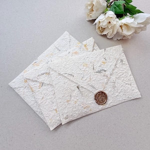 Könnte beinhalten: Drei handgefertigte, strukturierte Papierumschläge mit einem dekorativen Wachssiegel. Die Umschläge sind cremefarben mit Gold- und Silberflecken. Ein Strauß weißer Rosen befindet sich im Hintergrund.
