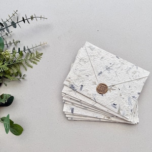 Könnte beinhalten: Ein Stapel handgefertigter Papierumschläge mit einer strukturierten, gesprenkelten Optik. Jeder Umschlag ist mit einem runden, bronzefarbenen Wachssiegel verschlossen. Das Papier hat blaue und graue Flecken. Grünzeug ist im Hintergrund zu sehen.