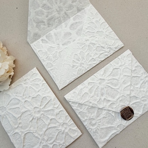 May include: Three white envelopes featuring a raised, embossed floral pattern across the entire surface. One envelope is open, one is closed, and one is sealed with a small, round, dark brown wax seal. The paper has a textured, fibrous appearance.