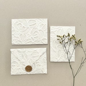 May include: Two rectangular off-white envelopes and one matching rectangular card, all featuring a raised, textured floral pattern. One envelope is sealed with a circular gold wax seal. A sprig of dried white flowers rests beside the items.