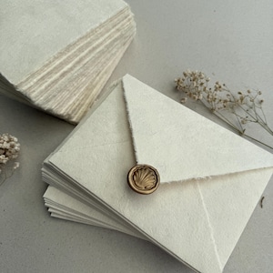 Op de afbeelding: Stapel crèmekleurige handgemaakte papieren enveloppen met gescheurde randen, verzegeld met een gouden schelpenzegel. Een tweede stapel bijpassend papier is zichtbaar op de achtergrond. Gedroogde bloemen zijn bij de enveloppen geplaatst.