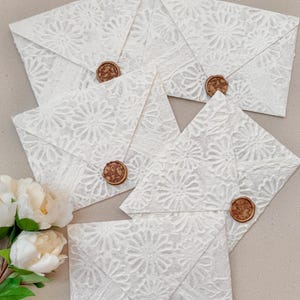 May include: Five white envelopes featuring a textured floral pattern and a circular bronze-colored wax seal on each flap. The envelopes are arranged in a cluster, and a small bunch of white peony flowers with green leaves is visible on the left side.