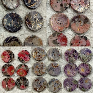 May include: A collection of decorative buttons with floral and abstract designs. The buttons feature a variety of colors, including pink, purple, gold, and blue. Some buttons have gold leaf accents. The buttons are round and have two or four holes for sewing.