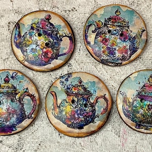 Puede incluir: Cinco botones redondos de madera con un diseño de tetera vintage. Cada botón presenta una colorida ilustración floral de tetera con una combinación de colores azul, morado y rosa. Los botones tienen una apariencia envejecida y desgastada.