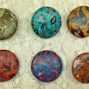 Könnte beinhalten: Sechs runde, dekorative, stoffbezogene Knöpfe mit Paisley-Mustern in verschiedenen Farben, darunter Türkis, Orange und Rot. Jeder Knopf ist von 1 bis 6 nummeriert.
