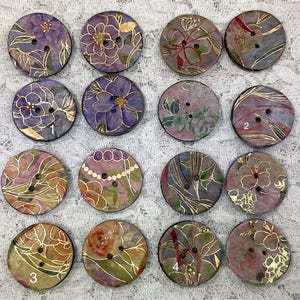 May include: Assortment of 12 round, wooden buttons with a variety of floral and abstract designs in shades of purple, pink, gold, and brown. The buttons have two holes each and are numbered 1, 2, 3, and 4.