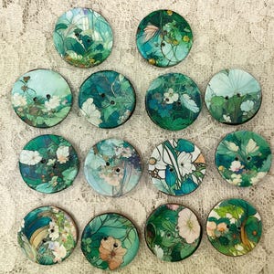 May include: A set of 14 round, green and blue buttons with floral and nature-inspired designs. The buttons are made of wood and have two holes each.
