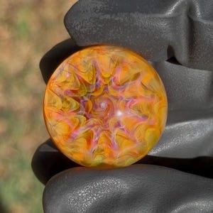 May include: A circular glass marble featuring a swirling pattern of vibrant orange, yellow, and magenta hues. The design radiates from a central point, creating a complex, layered effect within the smooth, polished, and translucent glass surface.