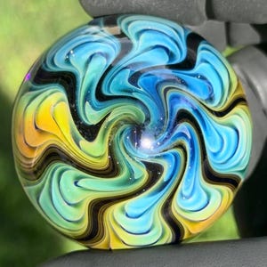 May include: A round glass marble featuring a swirling pattern of blue, teal, yellow, and black colors. The design consists of fluid, interconnected waves that radiate from the center, creating a smooth, polished surface with a glossy finish.